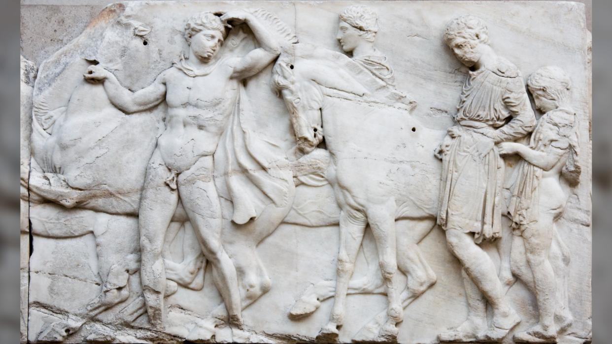  The Elgin Marbles stolen from Greece. From left to right we see a horse, a naked Greek man leading the way, whilst looking back at the others. Then there's another horse. Next to the horse is a person, and behind the horse if a person clothed in a tunic and a child with cloth draped around their shoulders. 