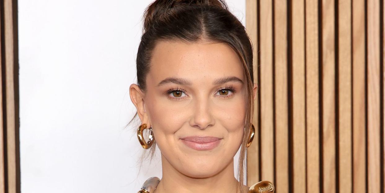 millie bobby brown, a young woman, wears her hair up and a black strapless gown with a sculpted metal neck piece and large hoop earings while posing for photographers at the glamour women of the year 2023 arrivals