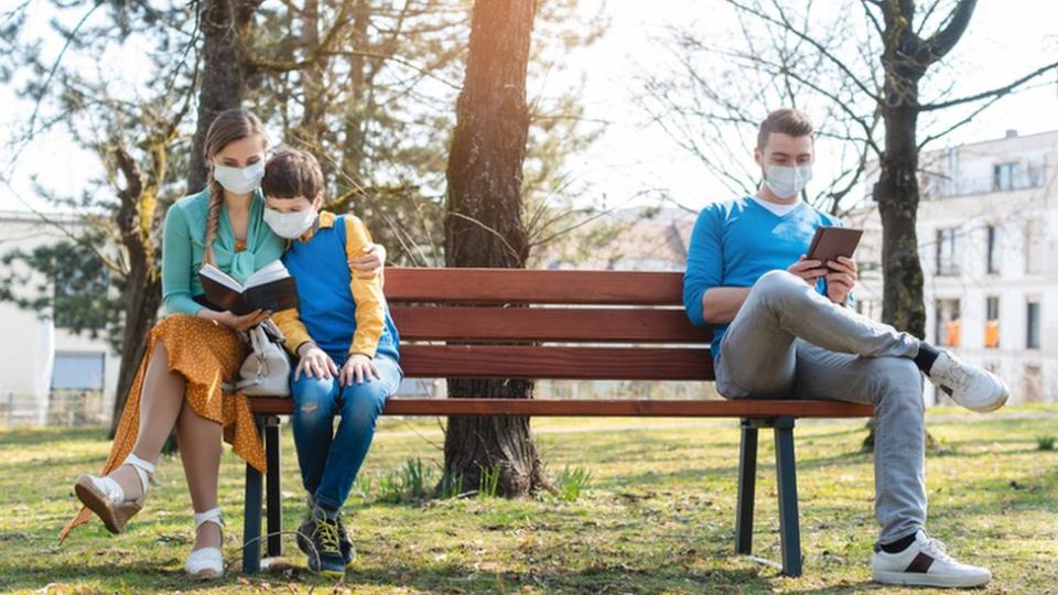 Una mujer con un niño y uno hombre sentados separados en un banco de plaza. Todos con mascarilla.