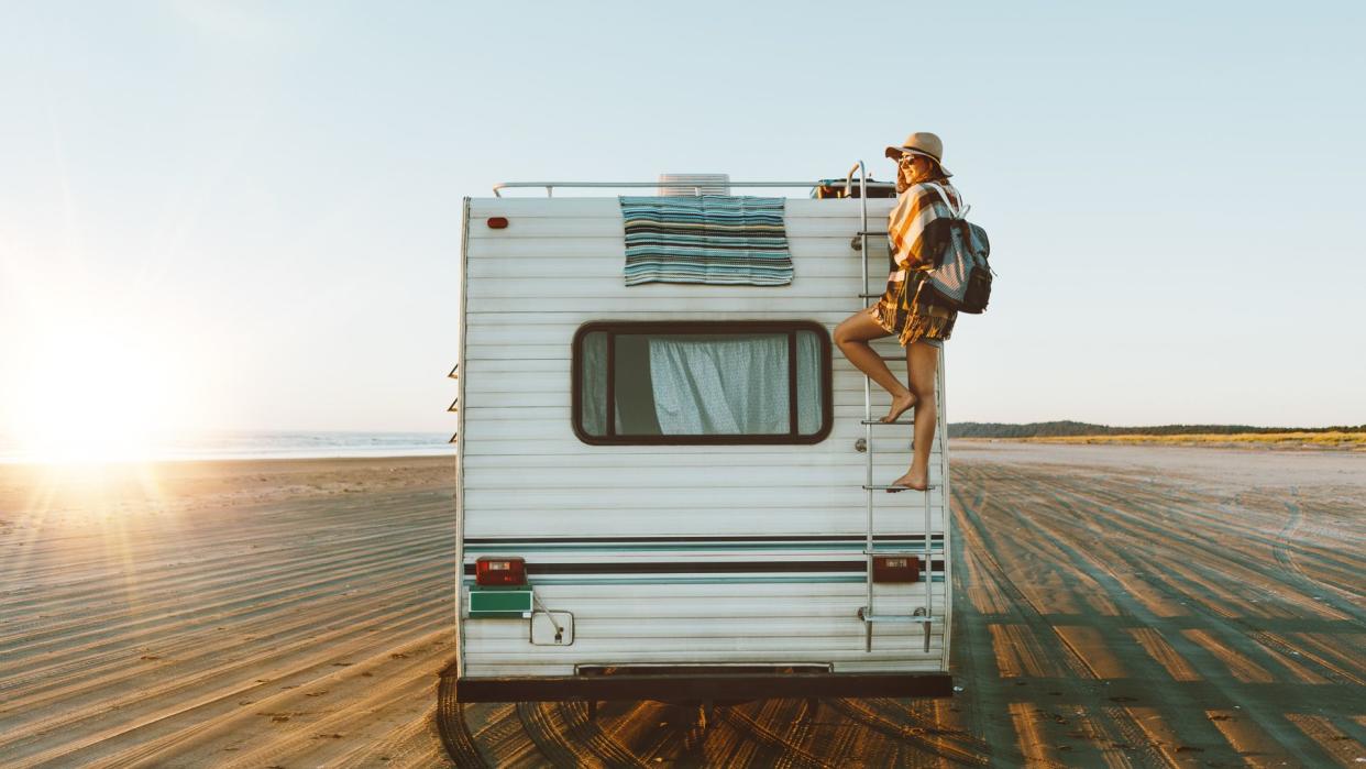 Charming young woman with nice smile with hat, sunglasses, backpack climing on recreational vehicle on the ocean beach at sunset.