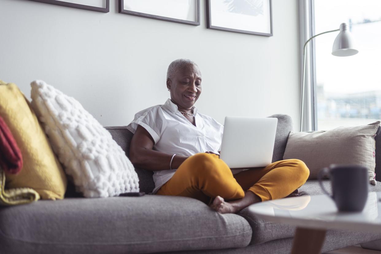 A black senior woman sits comfortably on her couch in her apartment. She is using a laptop and is consulting from home during the COVID-19 pandemic.