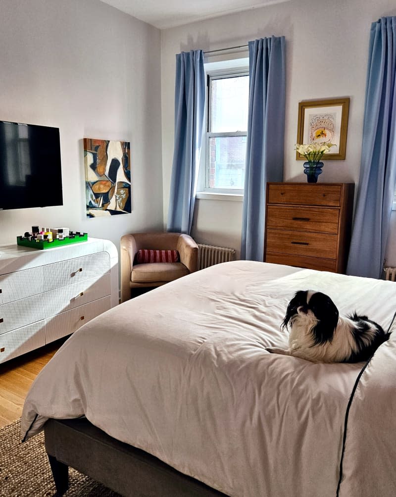 white bedroom with light blue curtains, sisal rug, curved white dresser, and dog on bed