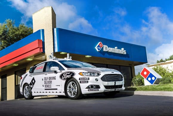 A white Ford Fusion sedan with large logos and visible self-driving hardware parked outside a Domino's pizza restaurant.