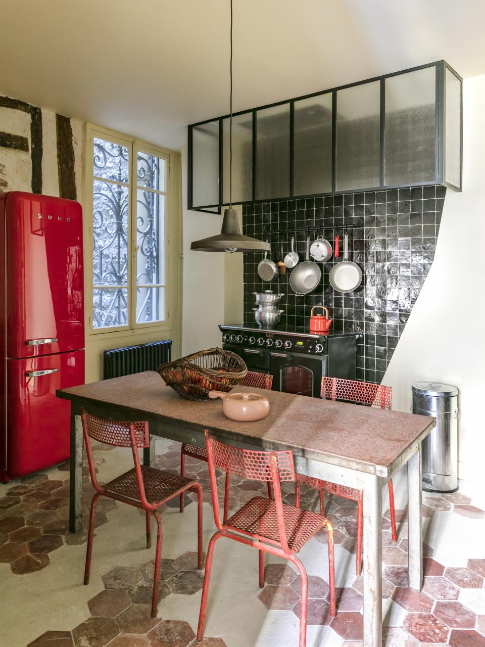 A cherry red Smeg refrigerator anchors the kitchen, in which all the pots and pans are available through Merci. A pink ceramic piece by Russel Wright can be seen on an industrial 1950s table. Around the table, vintage metal bistro chairs from the same decade have been placed.