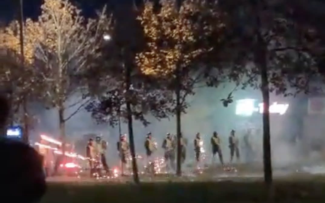 A line of police officers stand surrounded by smoke behing trees