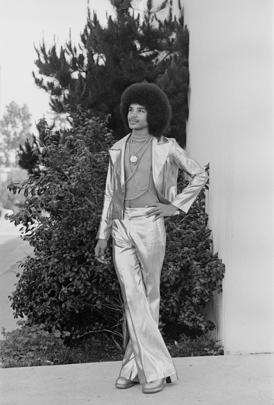 A person wearing a shiny, metallic outfit with flared pants and layered necklaces poses confidently outdoors