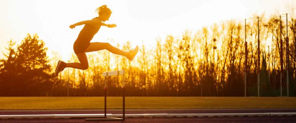 Athletic woman jumping above the hurdle on stadium running track during sunset