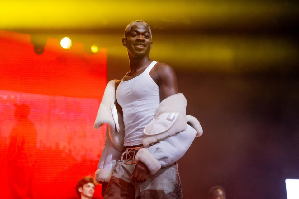 Lil Nas X in tank top and unique fluffy shoulder accessory on stage with red background