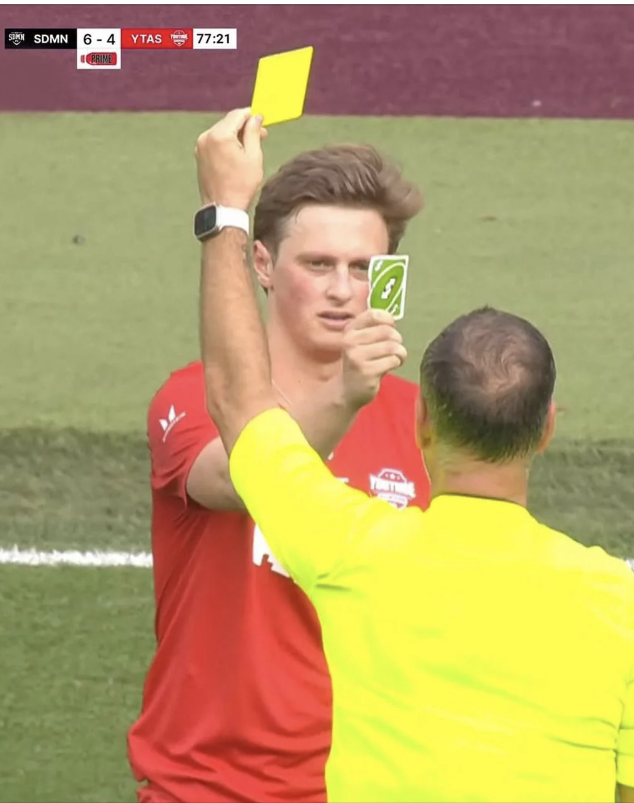 A soccer referee gives a yellow card while a player amusingly displays a green Uno reverse card. Scoreboard shows SDMN 6-4 YTAS with 77:21 on the clock