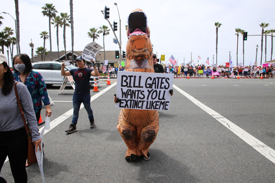 HUNTINGTON BEACH, CALIFORNIA, UNITED STATES - 2020/05/09: A protester wearing a dinosaur costume holds a placard that says Bill Gates Wants You Extinct Like Me during the demonstration. Citizens staged a protest in front of the Huntington Beach Pier to demand for the reopening of the California economy. (Photo by Stanton Sharpe/SOPA Images/LightRocket via Getty Images)