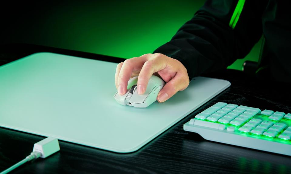 Um mouse para jogos Razer Viper V3 Pro branco é colocado em cima de um tapete de mouse branco em uma mesa preta, com um teclado branco brilhando com iluminação verde situado ao seu lado.