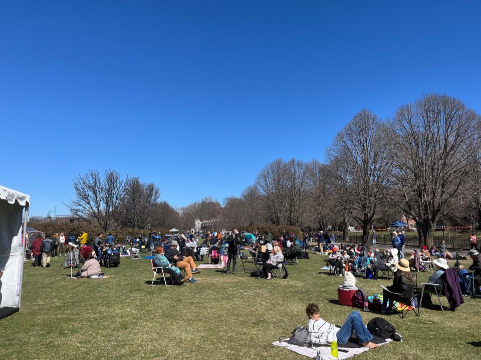 A grassy field with a crowd of people lounging under a blue sky.