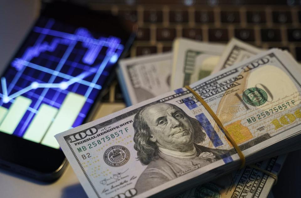 A stack of $100 bills next to a smartphone display showing a stock chart.