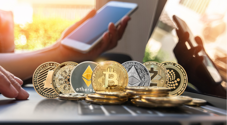 hot cryptos to buy: Various cryptocurrency coins are on a computer in front of someone holding a phone