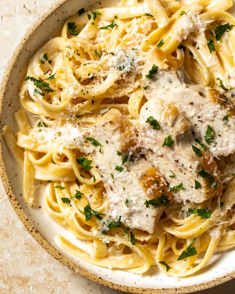 An overhead photo of a bowl of chicken fettucine Alfredo with grated parmesan cheese and parsley on top on a tan stone background.