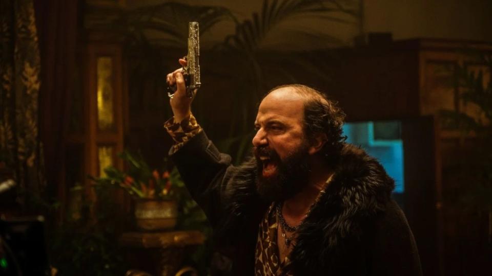 A balding man dressed in furs and gaudy jewelry holds up a gun and screams