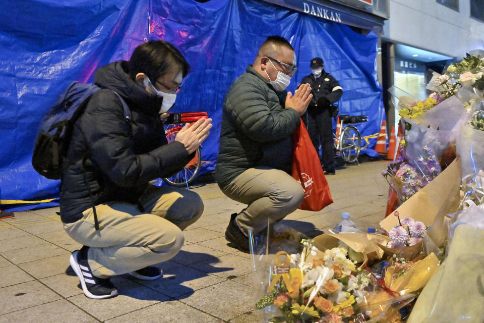 Mourners pray in front of offerings near a building, background, where a fire broke out, in Osaka, western Japan Thursday, Dec. 30, 2021. The suspect in the fire that killed 25 people died Thursday at a hospital where he was being treated for burns and smoke inhalation, police said Friday. (Kyodo News via AP)
