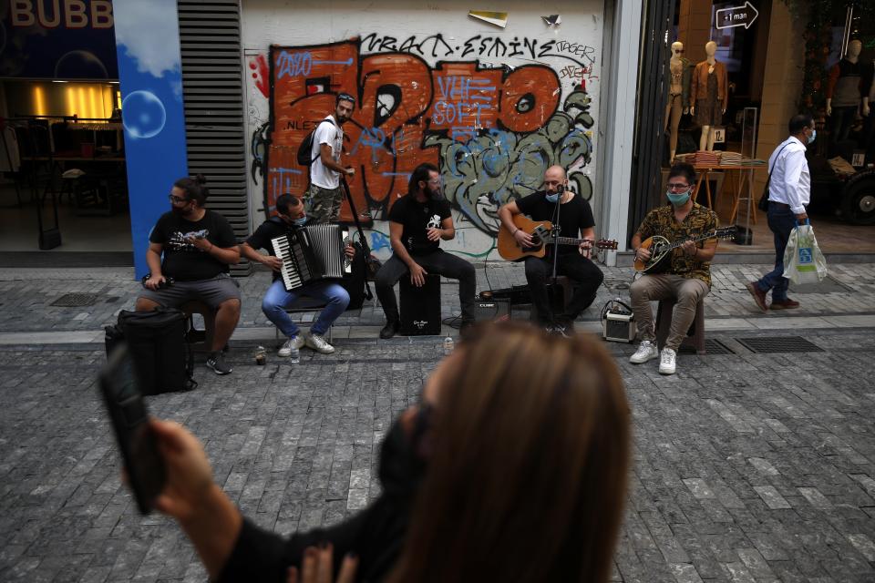 Musicians wearing face masks to prevent the spread of the coronavirus, perform as a woman records a video with her cellphone on Ermou Street, Athens' main shopping area, Monday, Oct. 26, 2020. Greece's government has imposed a nightly curfew in greater Athens and other areas with high infection rates as well as more generalized mask use as the daily number of deaths and infections nationwide has rising in recent weeks. (AP Photo/Thanassis Stavrakis)