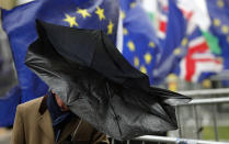 A man shelters with an umbrella as he walks by Pro EU protestors holding flags opposite the Houses of Parliament in London, Thursday, April 4, 2019. The British government and senior opposition figures were meeting Thursday in search of a new plan on how the country leaves the European Union as Prime Minister Theresa May tried to stop her shift toward compromise from splitting her Conservative Party. (AP Photo/Frank Augstein)