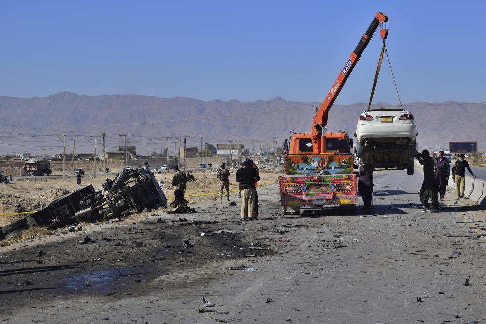 Workers remove a damage car while security officials inspect the site of a suicide bombing in Quetta, Pakistan, Wednesday, Nov. 30, 2022. A suicide bomber blew himself up near a truck carrying police officers on their way to protect polio workers outside Quetta, wounding more than 20 others, officials said. (AP Photo/Arshad Butt)