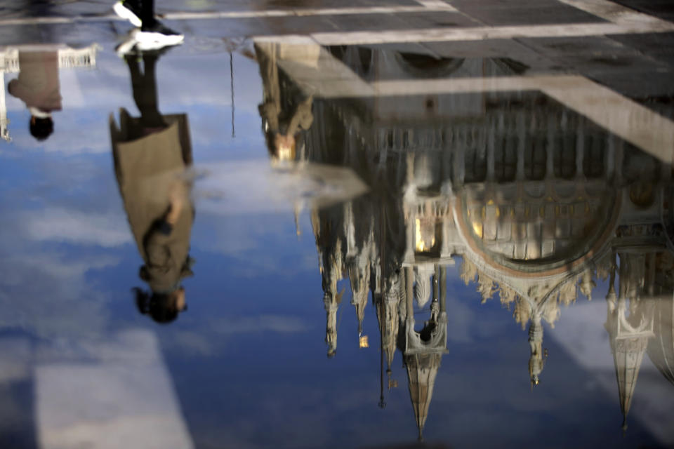 A tourist is reflected in a puddle along with St. Mark cathedral, as water starts rising again in Venice, Italy, Saturday, Nov. 16, 2019. High tidal waters returned to Venice on Saturday, four days after the city experienced its worst flooding in 50 years. (AP Photo/Luca Bruno)