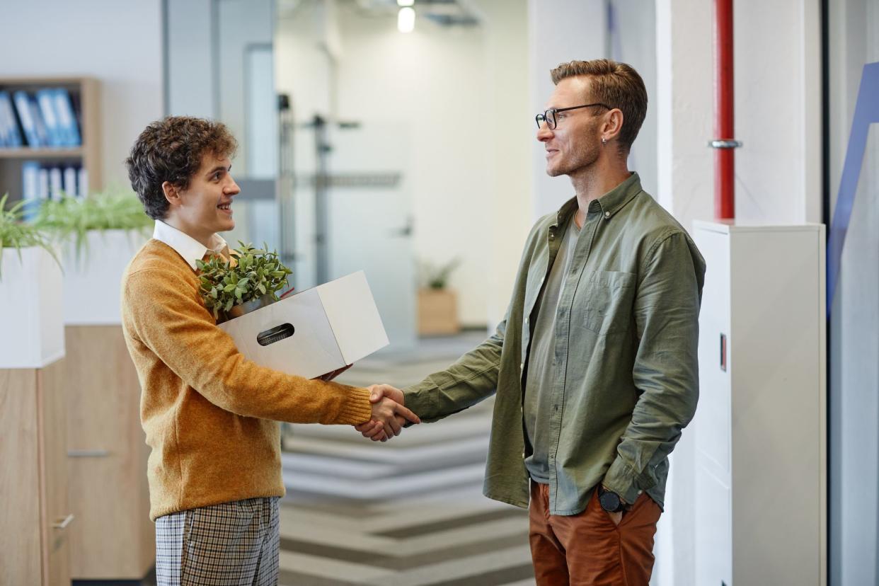 Side view portrait of new employee shaking hands with colleagues while holding box with personal belongings