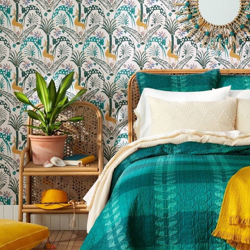 A bohemian bedroom setup with a rattan bed and side table, and a patterned wallpaper. Decor includes a plant, mirror, and textiles