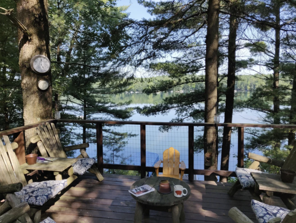 A scenic view from a wooden deck overlooking a lake surrounded by trees, with Adirondack chairs and a small table