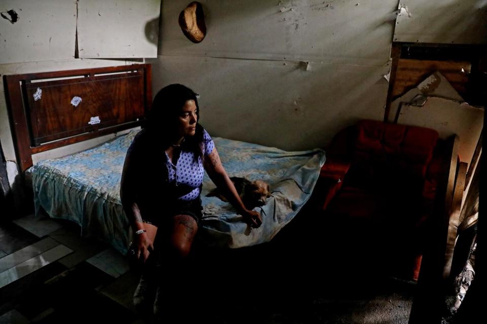 A woman with dark hair, in a pale top and dark shorts, sitting on a bed