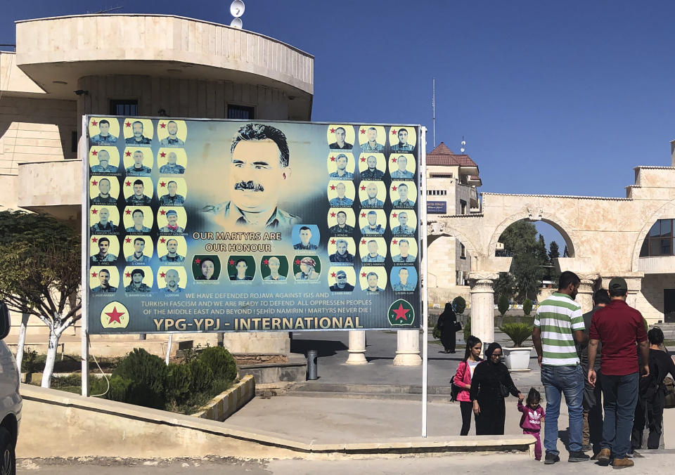 People pass a giant banner with a portrait of jailed leader of the Kurdistan Workers' Party, or PKK, Abdullah Ocalan, center, with other portraits of killed Kurdish fighters, set at the border crossing between Iraq and Syria's Kurdish-held, in Semelka, northeast Syria, Monday Oct. 14, 2019. (AP Photo/Hussein Malla)