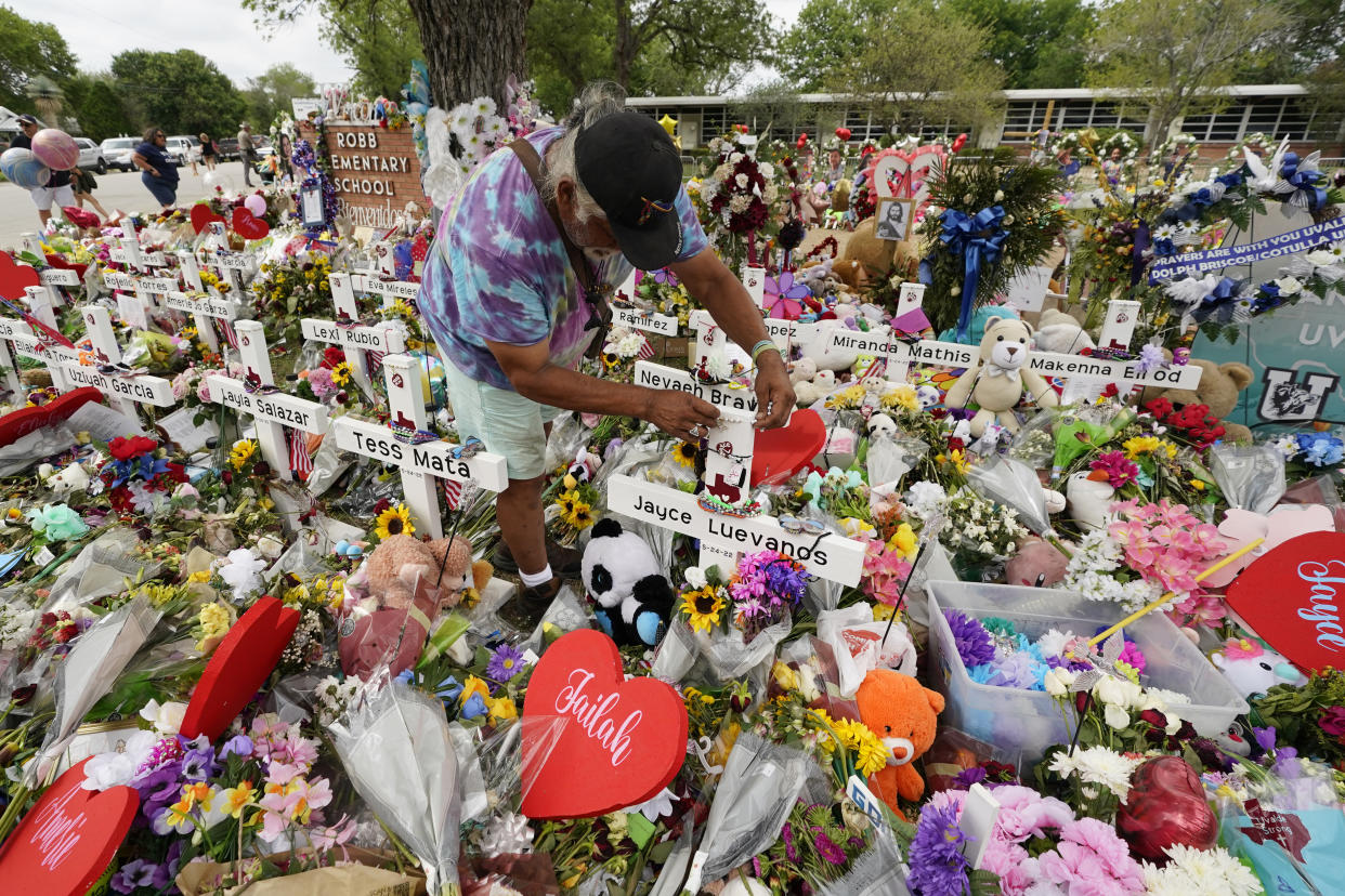 A visitor places stands among crosses, flowers and stuffed animals. The Robb Elementary School sign and building are seen in the background.