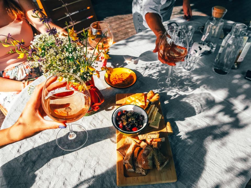 An image of an outdoor table with a plate of olives and bread, surrounding by people toasting with glasses of wine.