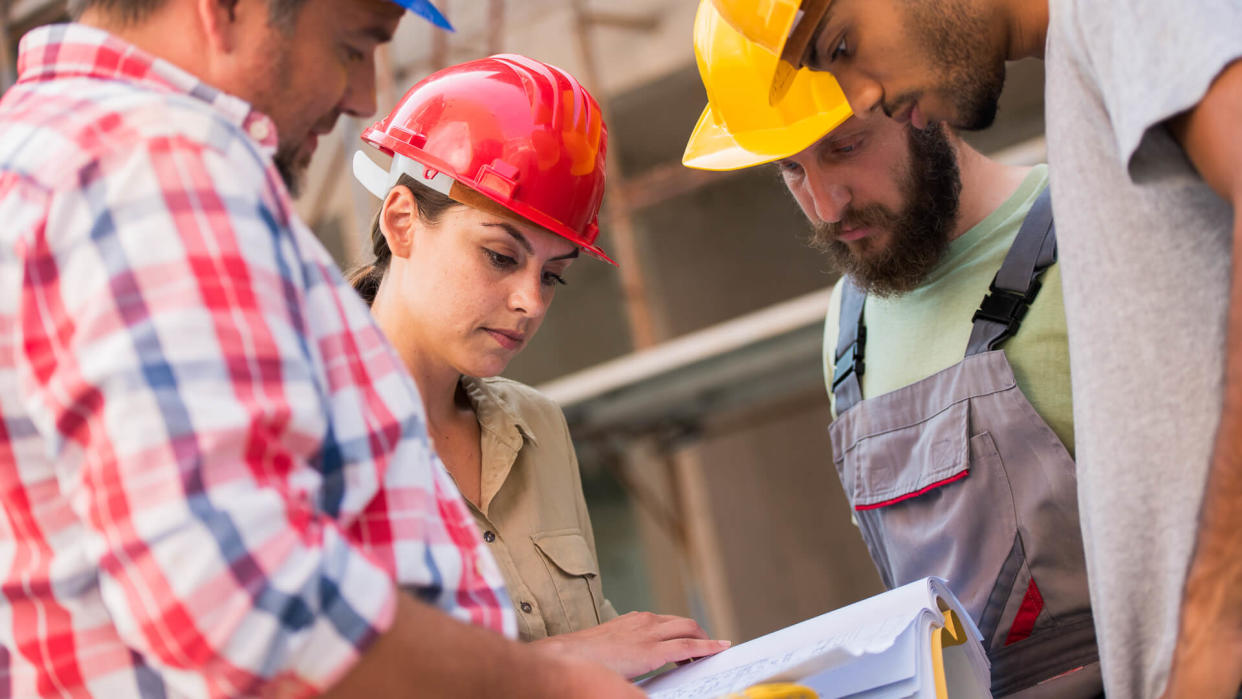 Three workers gathered in a circle around an architect holding blueprints