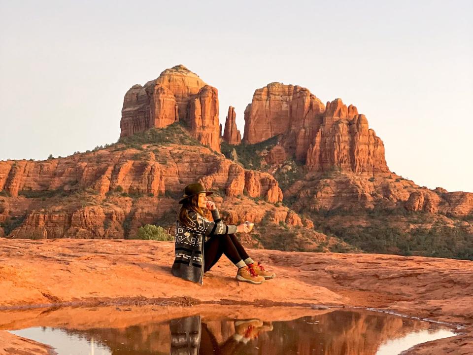 Emily sits on the ground facing to the right. Behind her are large red-rock formations, and in front of her are a small body of water and a cactus.