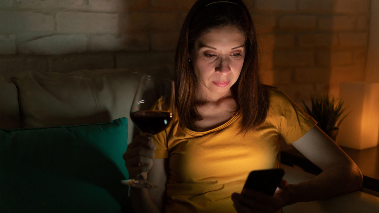  woman drinking a glass of wine at night as she looks at her smartphone 