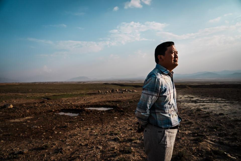 A man in a blue plaid shirt and khaki pants stands amid barren land under blue skies