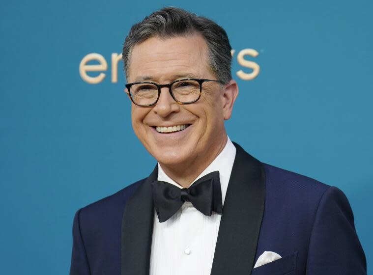 Stephen Colbert in glasses and a blue tuxedo smiling and standing in front of a blue-green background