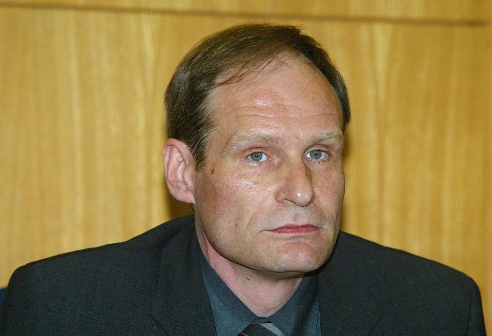 A man in a suit and tie sits against a wood-paneled background. Name of the person is unknown