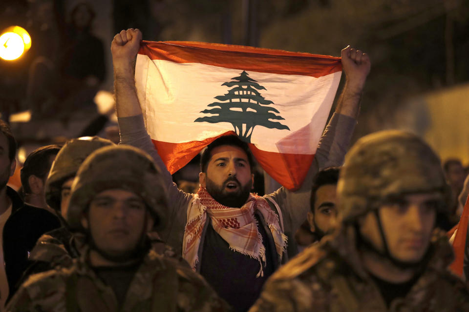 An anti-government protester holds his nation flag, as he stands behind Lebanese army soldiers during a clash between the anti-government protesters and the supporters of the Shiite Hezbollah and Amal Movement groups, in Beirut, Lebanon, early Monday, Nov. 25, 2019. Security forces fired tear gas amid confrontations in central Beirut that went into Monday morning between Hezbollah supporters and demonstrators protesting against Lebanon's political elite. (AP Photo/Hussein Malla)