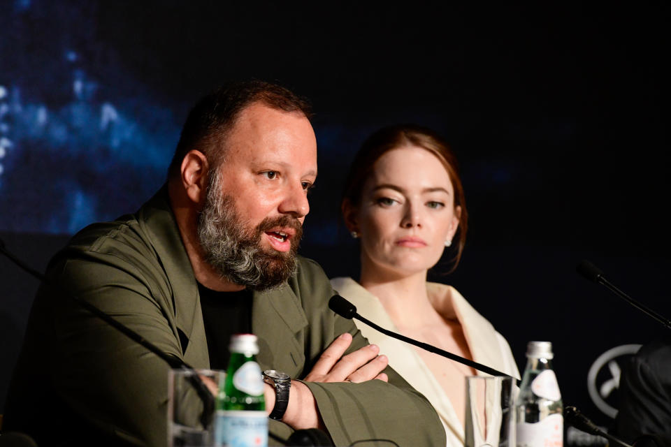 Yorgos Lanthimos and Emma Stone at a press conference table, Yorgos speaking into a microphone, and Emma attentively listening