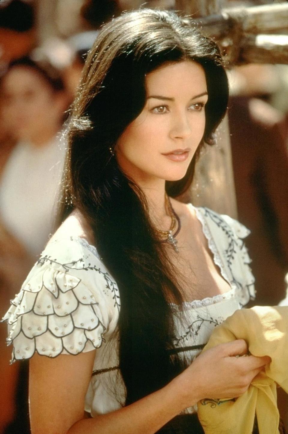 Catherine Zeta-Jones in a period film costume with intricate sleeve details and long, flowing hair. She is holding a beige cloth, with a neutral expression