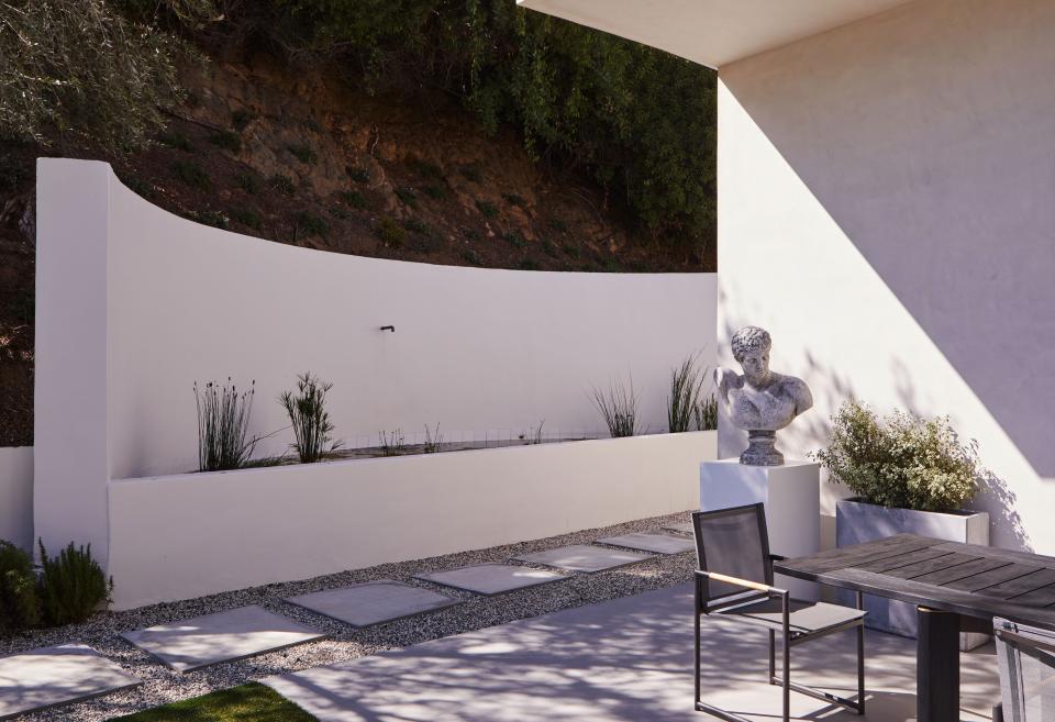 In this exterior area, adorned with a cast concrete sculpture of a man placed on a custom white pedestal, the Sonoma forge rustic nickel spout is from EuroConcepts and the Ora dining table and chairs are from Harbour Outdoor.