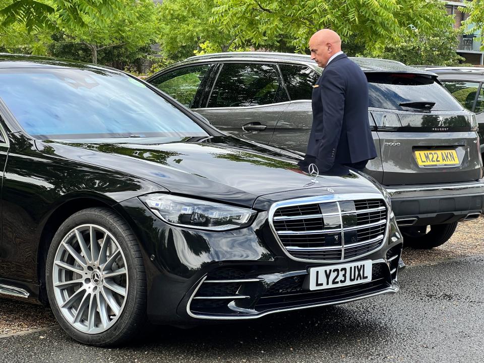 A man in a navy suit walks around a black Mercedes Benz S-Class