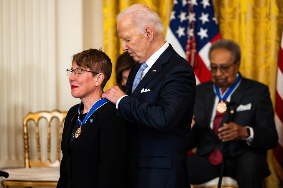 a woman wearing a nasa pin smiles with red lipstick while an old man in a suit hangs a medal necklace around her neck behind her.