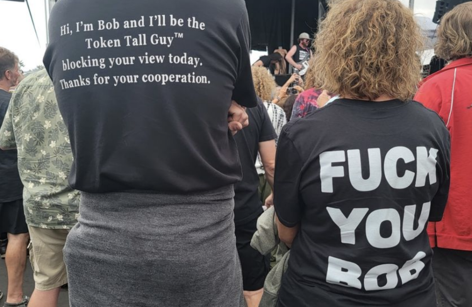 Tall guy with back of T-shirt saying "Hi, I'm Bob and I'll be the Token Tall Guy blocking your view today, thanks for your cooperation," and the woman with "Fuck you Bob" on the back of her shirt