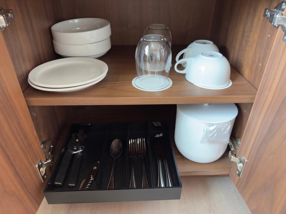 Glass plates, cups, and mugs above a tray of silverware in a wooden cabinet