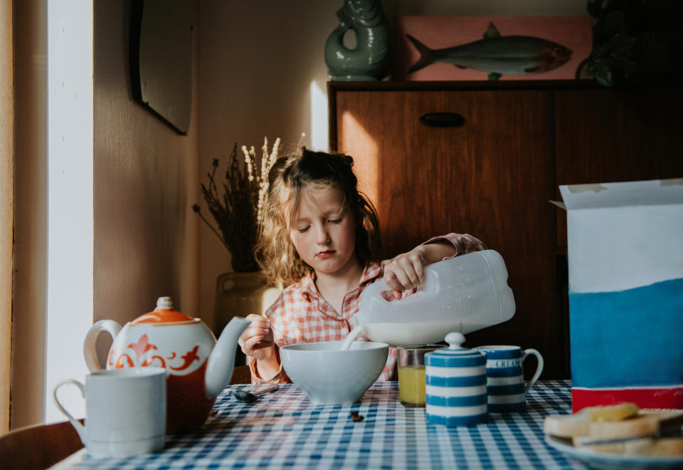 A cute young girl wearing pyjamas sits at a breakfast table in the morning. She looks sleepy as she pours milk over her cereal.