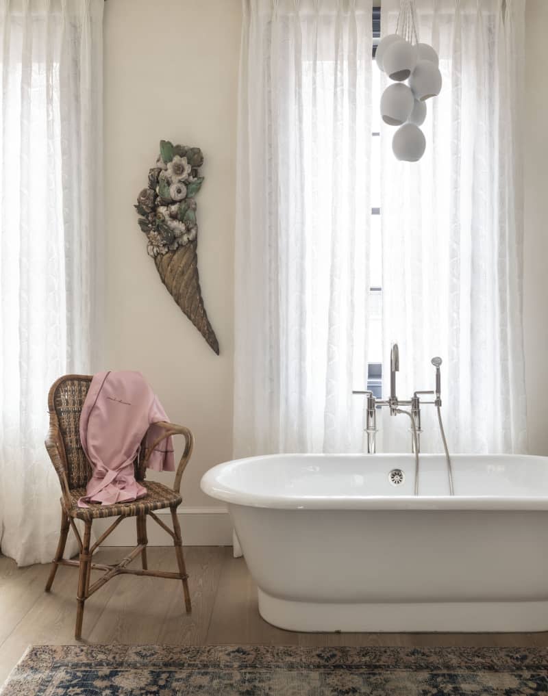 A wicker chair next to a white tub across from two large windows.