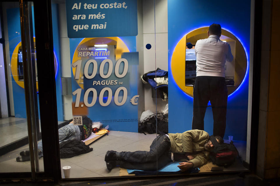 A man uses a cash dispenser to withdraw money as homeless men sleep inside a bank in Barcelona.