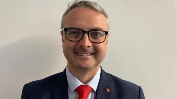 Juan Watterson looking directly into the camera, smiling. He is wearing a navy blue jacket, pale blue shirt and bright red tie, along with black-rimmed glasses. He has a red pin badge featuring a yellow three-legs-of-Mann symbol in the buttonhole of his jacket.